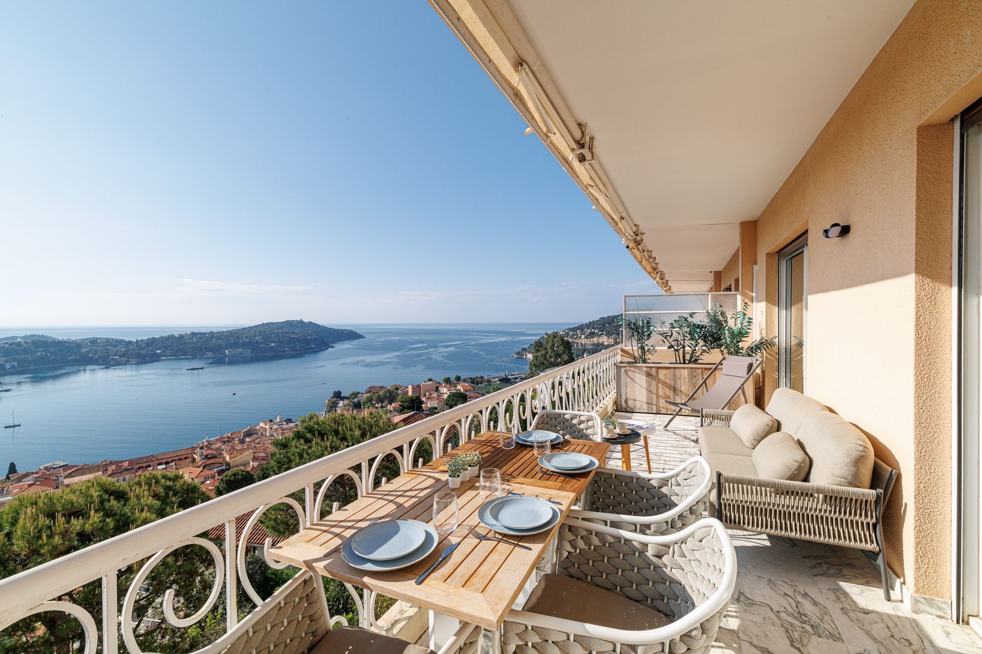 Balcony with dining table overlooking a scenic coastal view with turquoise sea and green hills.